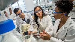 Three CELS students wearing lab safety glasses and coats talking in front of a microscope